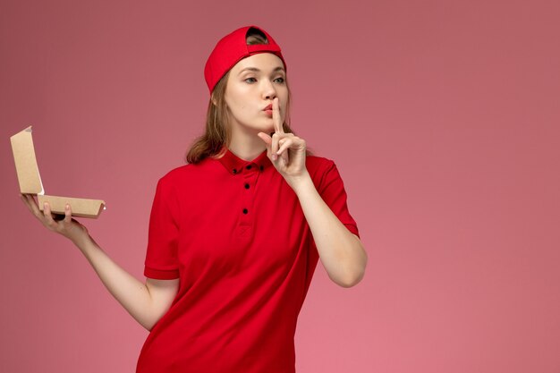 Front view female courier in red uniform and cape holding little delivery food package on pink floor delivery service company uniform work