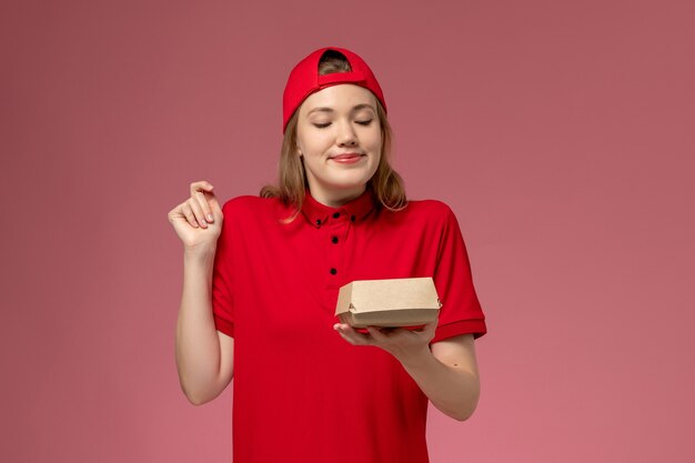 Front view female courier in red uniform and cape holding little delivery food package on pink wall, delivery job service company uniform work girl