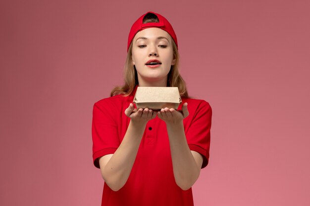 Front view female courier in red uniform and cape holding little delivery food package on pink wall, delivery job service company uniform work