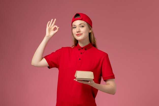 ピンクの壁に小さな配達食品パッケージを保持している赤い制服と岬の正面図の女性の宅配便、配達サービス会社の制服の労働者