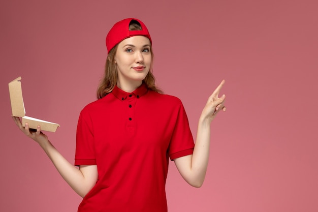 Front view female courier in red uniform and cape holding little delivery food package on the pink wall, delivery service company uniform job worker