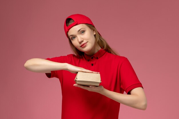 Вид спереди женщина-курьер в красной униформе и накидке с маленьким пакетом еды для доставки на розовой стене, работник службы доставки