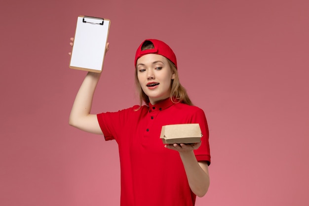 赤いユニフォームとピンクのデスクデリバリーサービス会社の仕事のユニフォームにメモ帳付きの小さな配達食品パッケージを保持している岬の正面図の女性の宅配便