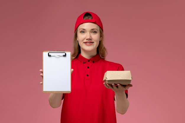 赤いユニフォームとピンクの壁にメモ帳付きの小さな配達食品パッケージを保持している岬の正面図の女性の宅配便、配達サービス会社の仕事の制服