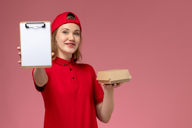 Front view female courier in red uniform and cape holding little delivery food package with notepad on pink wall, delivery service uniform girl job work
