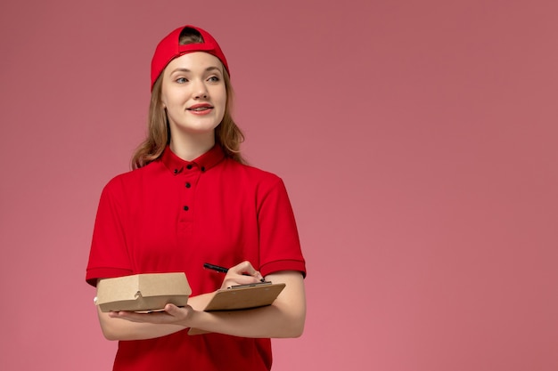 Front view female courier in red uniform and cape holding little delivery food package with notepad on the pink wall, delivery service uniform girl job worker work