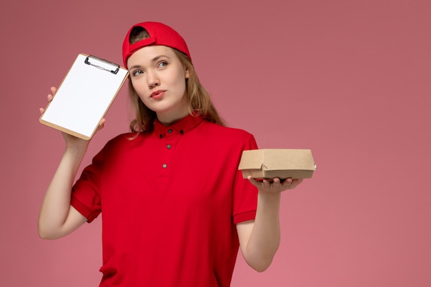 Вид спереди женщина-курьер в красной форме и накидке, держащая небольшой пакет продуктов для доставки с блокнотом на розовой стене, униформа службы доставки