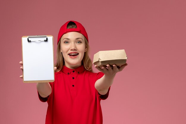 Front view female courier in red uniform and cape holding little delivery food package with notepad on pink wall, delivery service uniform girl work