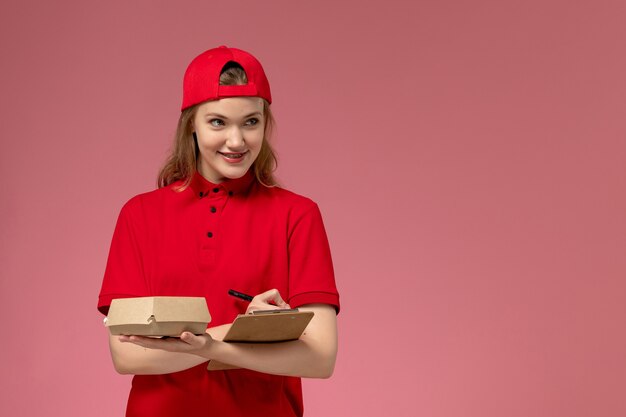 Front view female courier in red uniform and cape holding little delivery food package with notepad on pink wall, service uniform delivery job worker