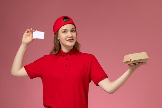 Corriere femminile di vista frontale in uniforme rossa e mantello che tiene piccolo pacchetto di cibo per la consegna con carta di plastica bianca sul muro rosa, consegna uniforme di lavoro di servizio