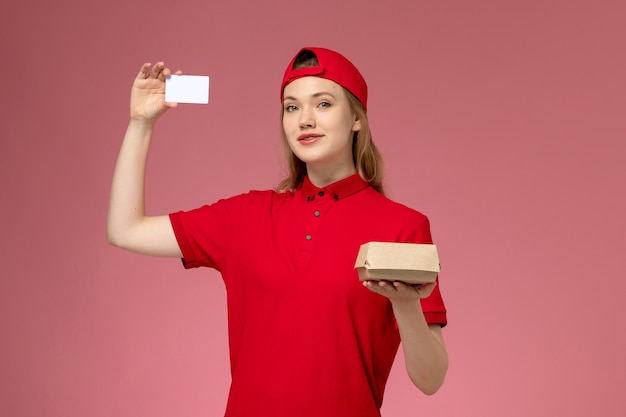ピンクの壁に白いプラスチックカードが付いた小さな配達食品パッケージを保持している赤い制服と岬の正面図の女性の宅配便、サービス制服配達の仕事