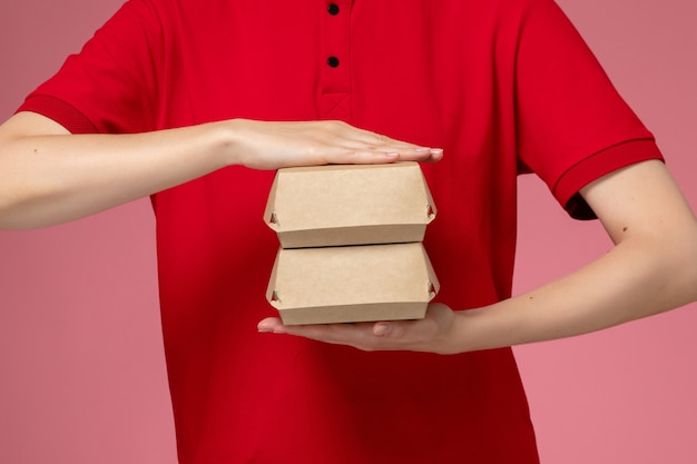 Front view female courier in red uniform and cape holding little delivery food packages on light pink wall, delivery service company uniform job worker