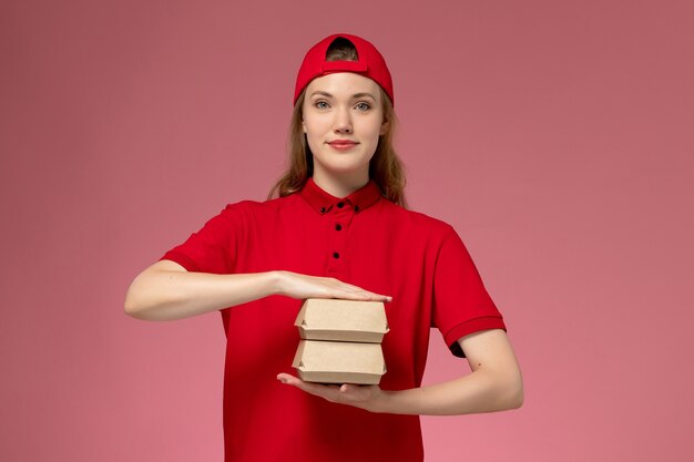 Front view female courier in red uniform and cape holding little delivery food packages on light pink wall, delivery service company uniform job