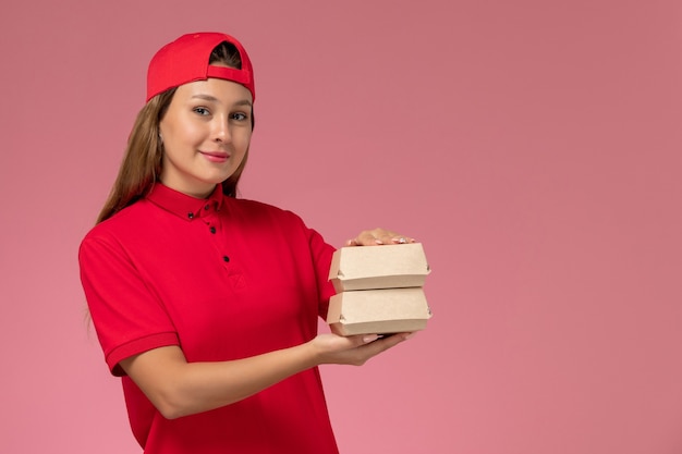 Вид спереди курьер-женщина в красной форме и накидке с маленькими пакетами еды для доставки на светло-розовой стене, единообразная служба доставки работы