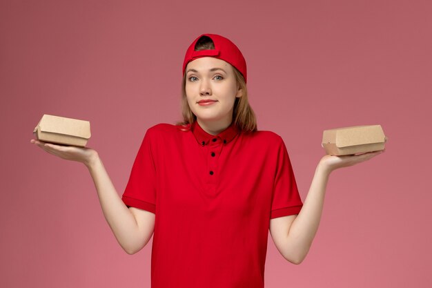 Front view female courier in red uniform and cape holding little delivery food packages on pink wall, worker delivery service company uniform job