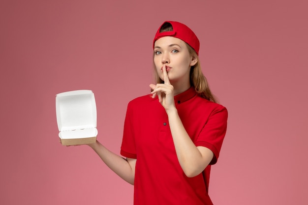 Вид спереди курьер-женщина в красной форме и накидке с маленьким пустым пакетом еды для доставки на розовой стене, работник службы доставки