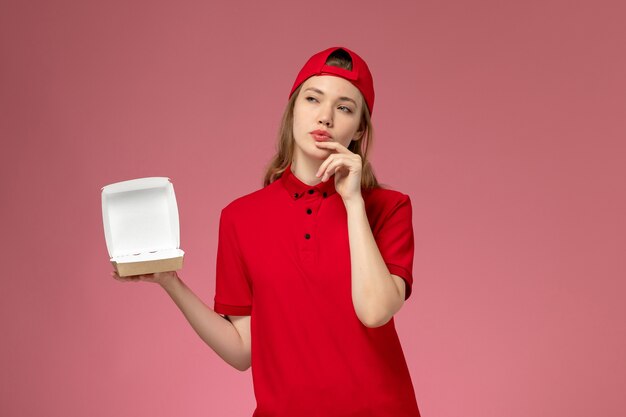 Front view female courier in red uniform and cape holding little empty delivery food package thinking on pink wall, delivery service company uniform job