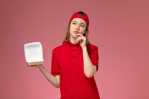 분홍색 벽, 배달 서비스 회사 유니폼 작업에 생각하는 작은 빈 배달 음식 패키지를 들고 빨간 유니폼과 케이프 전면보기 여성 택배