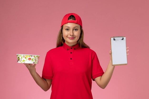 Front view female courier in red uniform and cape holding notepad and delivery bowl on pink background uniform job delivery service