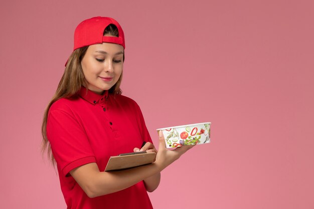 Front view female courier in red uniform and cape holding notepad and delivery bowl on pink background uniform worker job delivery service