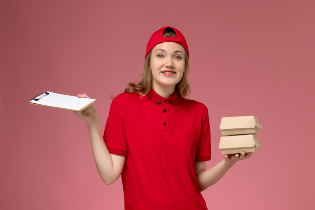 Front view female courier in red uniform and cape holding notepad and little delivery food packages on light-pink background service uniform delivery