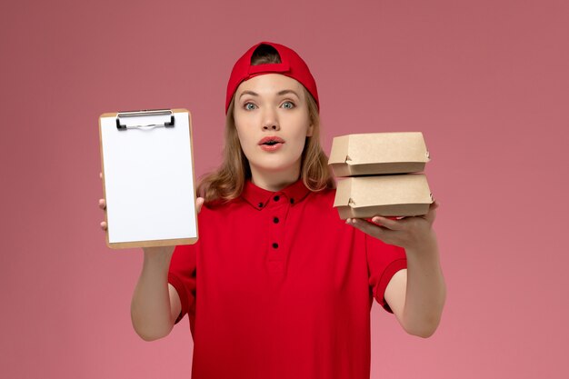 Front view female courier in red uniform and cape holding notepad and little delivery food packages on the light-pink wall, desk service uniform delivery