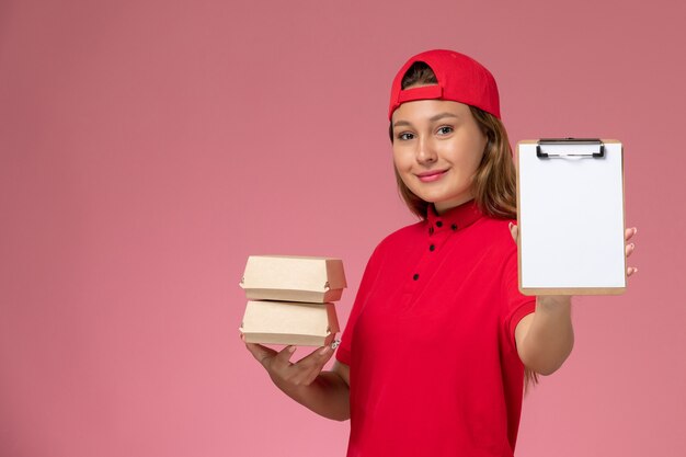 赤い制服とケープを保持しているメモ帳とピンクの壁に小さな配達食品パッケージの正面図の女性の宅配便、均一な配達サービス作業