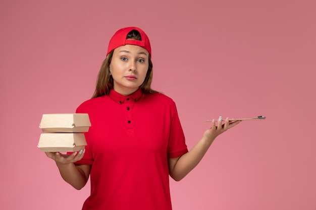 Free photo front view female courier in red uniform and cape holding notepad and little delivery food packages on the pink wall, uniform delivery service