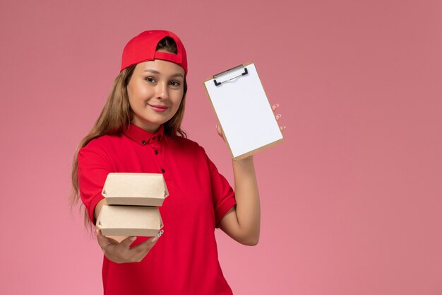 Front view female courier in red uniform and cape holding notepad and little delivery food packages on pink wall, uniform job delivery service