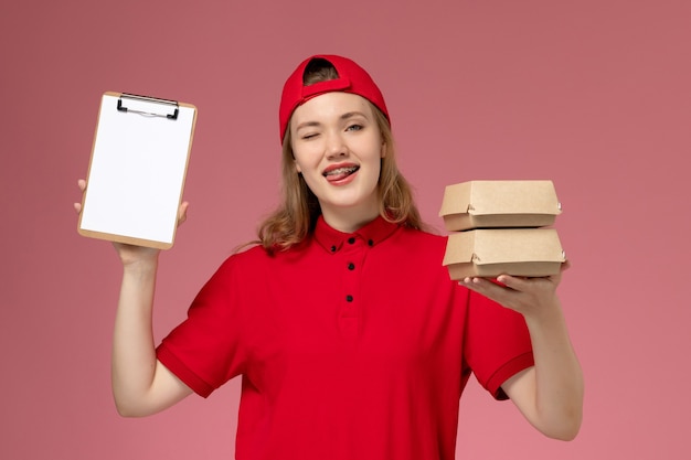 Front view female courier in red uniform and cape holding notepad and little delivery food packages winking on light-pink wall, service uniform delivery