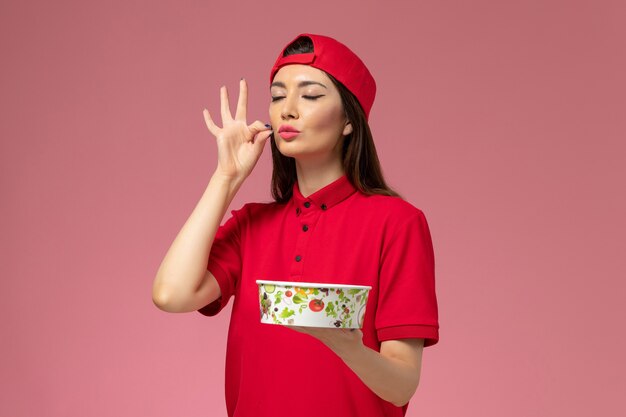 Front view female courier in red uniform cape with delivery bowl on her hands on light pink wall, uniform delivery employee