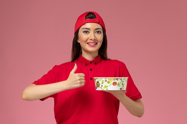 Front view female courier in red uniform cape with delivery bowl on her hands smiling on light pink wall, service delivery employee