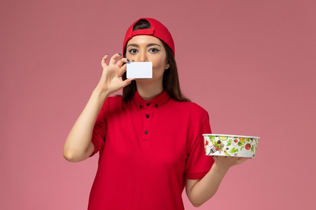 Курьер-женщина, вид спереди в красной форменной накидке с миской для доставки и белой карточкой на руках на светло-розовой стене, работа сотрудника по доставке униформы