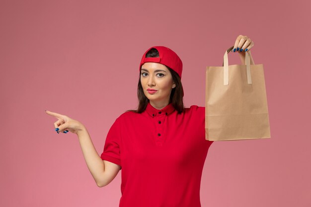 Front view female courier in red uniform cape with delivery paper package on her hands on pink wall, worker uniform delivery employee job