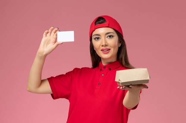밝은 분홍색 벽, 서비스 작업 배달 직원에 그녀의 손에 작은 배달 음식 패키지와 카드가있는 빨간색 유니폼 케이프의 전면보기 여성 택배