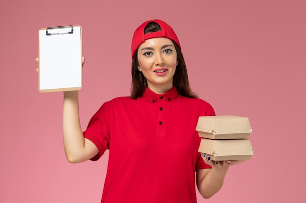 Front view female courier in red uniform cape with little delivery food package and notepad on her hands on light-pink wall, service delivery employee work