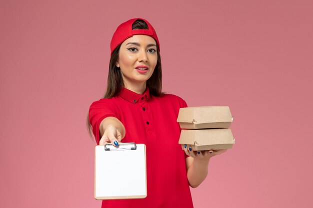 Женщина-курьер в красной униформе, вид спереди с небольшими пакетами еды для доставки и блокнотом в руках на светло-розовой стене, работа сотрудника службы доставки