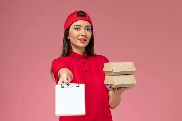 Front view female courier in red uniform cape with little delivery food packages and notepad on her hands on light-pink wall, service delivery employee work