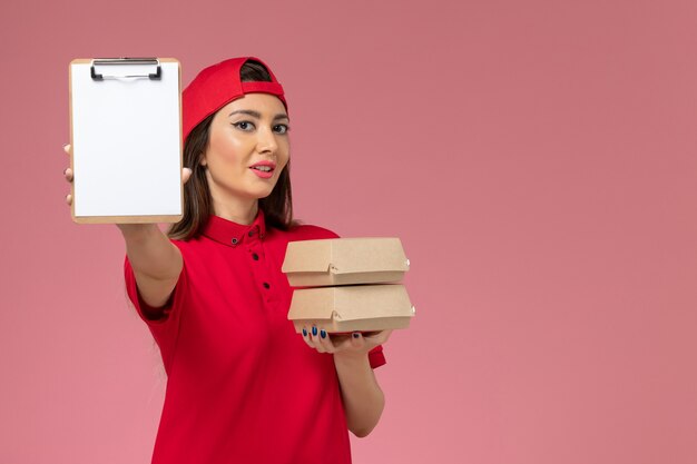 Free photo front view female courier in red uniform cape with notepad and little delivery food packages on her hands on light-pink wall, job service delivery employee