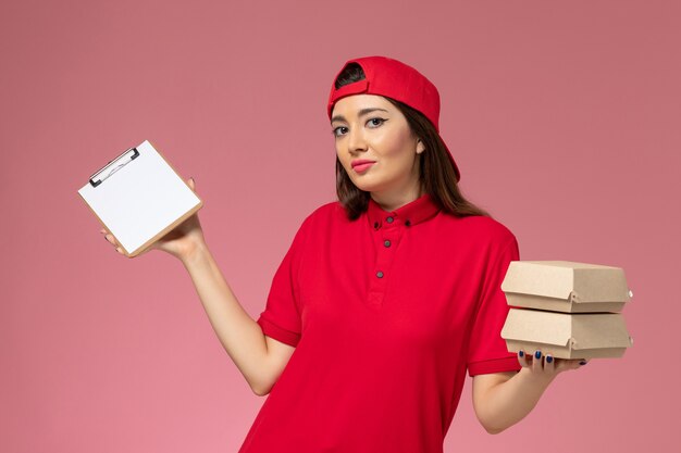 Front view female courier in red uniform cape with notepad and little delivery food packages on her hands on light-pink wall, service delivery employee work