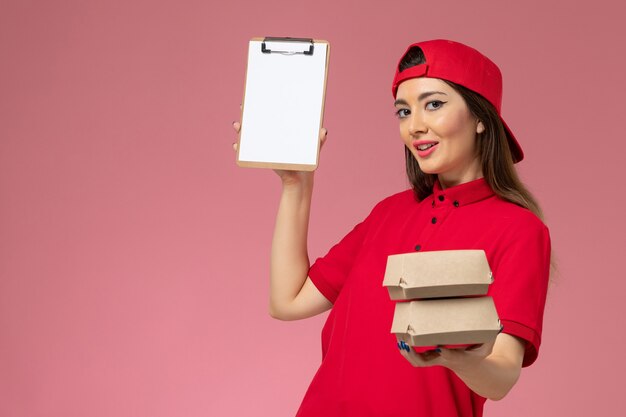 Front view female courier in red uniform cape with notepad and little delivery food packages on her hands on light-pink wall, work service delivery employee