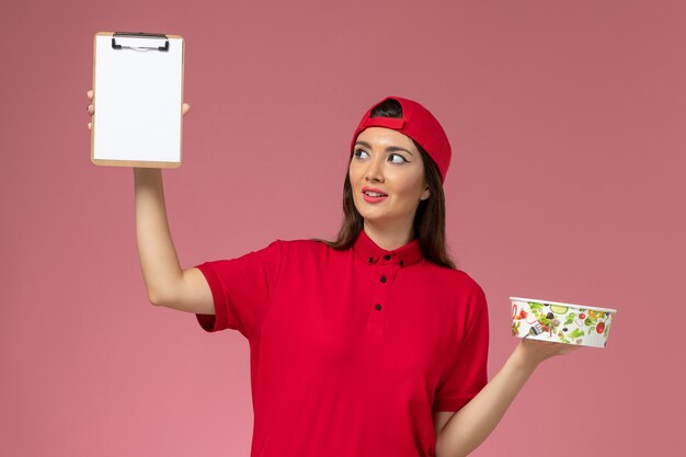 Front view female courier in red uniform cape with round delivery bowl notepad on her hands on light pink wall, uniform delivery employee work