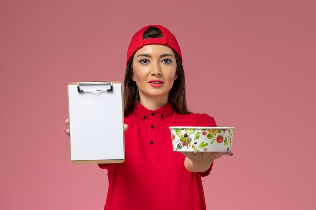 Front view female courier in red uniform cape with round delivery bowl and notepad on her hands on the light pink wall, uniform delivery employee