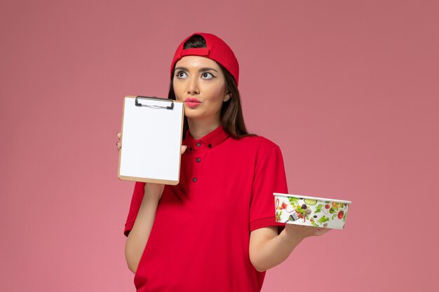 Front view female courier in red uniform cape with round delivery bowl and notepad on her hands thinking on the light pink wall, uniform delivery employee