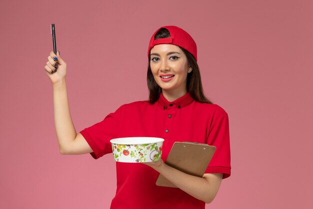 둥근 배달 그릇 메모장과 밝은 분홍색 벽에 그녀의 손에 펜, 균일 한 배달 직원 노동자와 빨간색 유니폼 케이프 전면보기 여성 택배