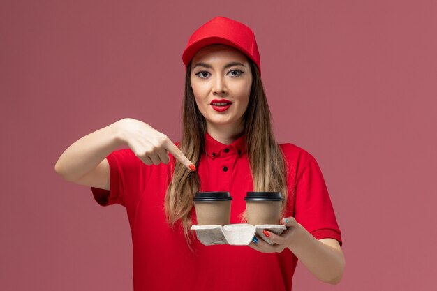 Front view female courier in red uniform holding brown delivery coffee cups on light-pink background service delivery uniform job