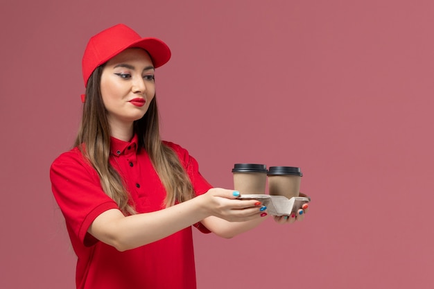 Corriere femminile di vista frontale in uniforme rossa che tiene le tazze di caffè di consegna marroni sul lavoro dell'uniforme di consegna del servizio di sfondo rosa chiaro