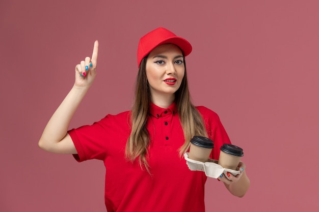 Front view female courier in red uniform holding brown delivery coffee cups on light-pink background service delivery uniform worker company