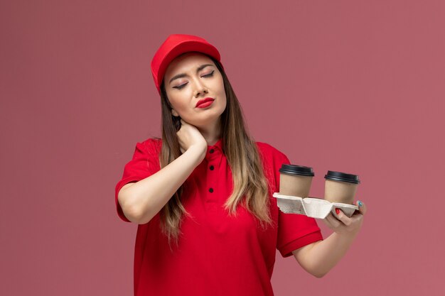 Front view female courier in red uniform holding brown delivery coffee cups on the light-pink background service delivery uniform worker job company