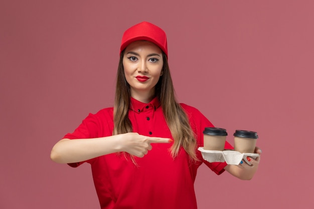 Front view female courier in red uniform holding brown delivery coffee cups on light-pink background service delivery uniform worker job female lady company